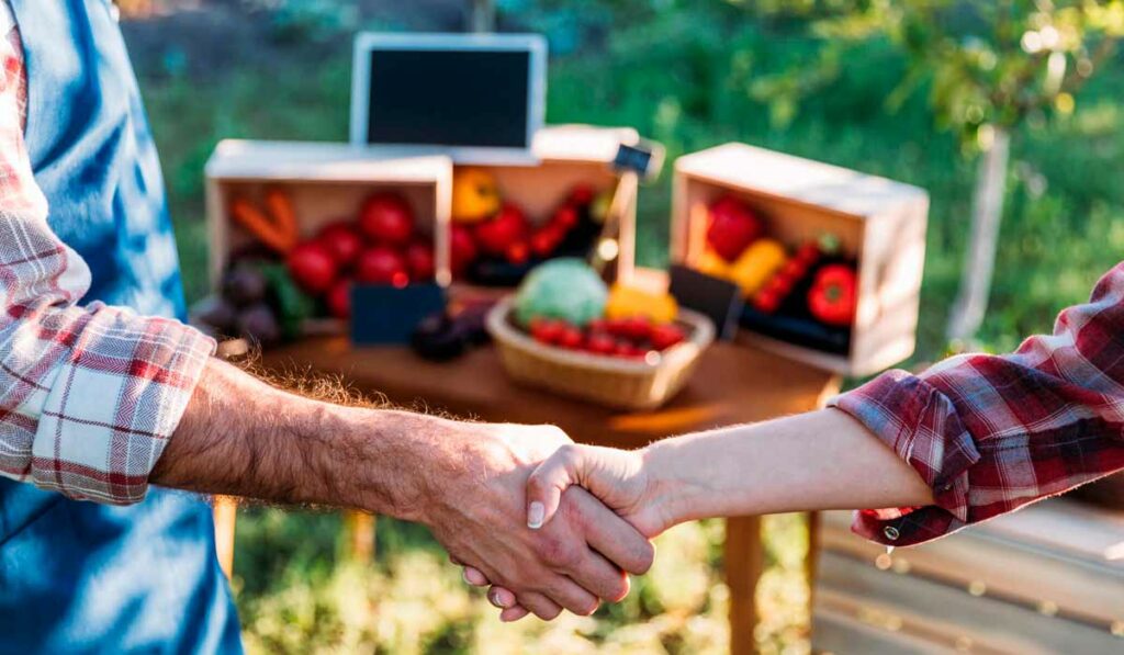 Agricultores concretizando um negócio com aperto de mãos e produtos agrícolas ao fundo