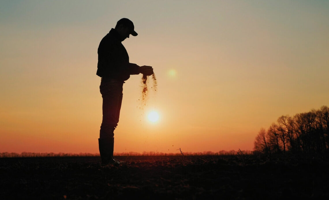 Agricultor deixando escorrer a terra das mãos com o por do sol ao fundo
