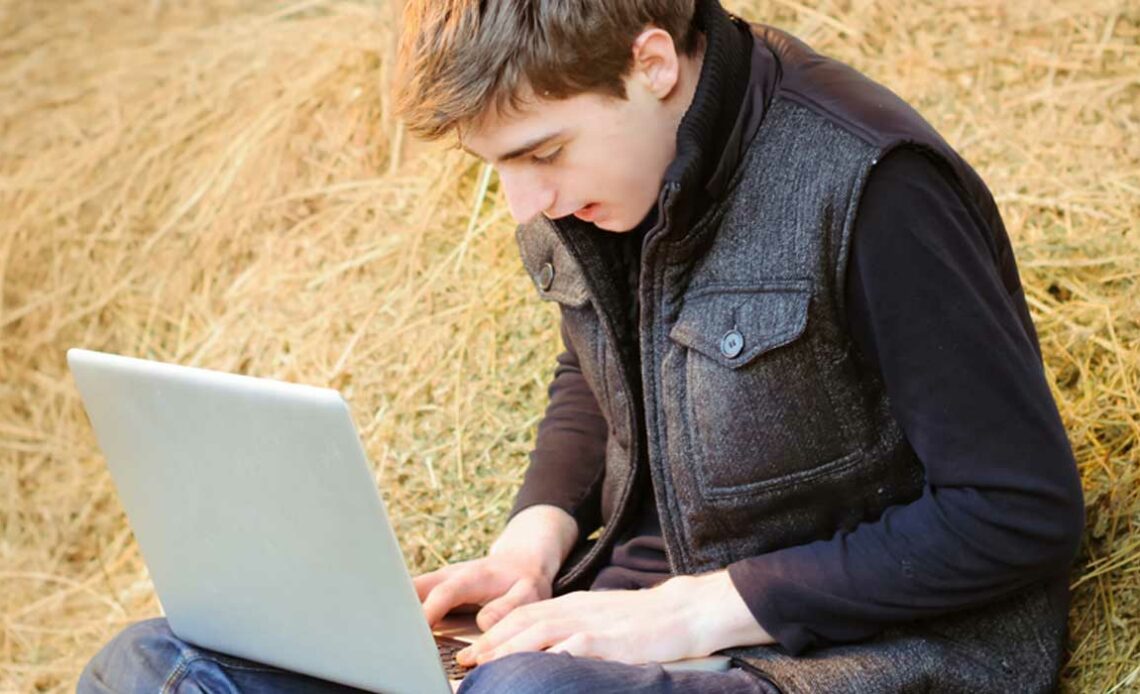 Jovem rural sentado no celeiro com feno e um laptop